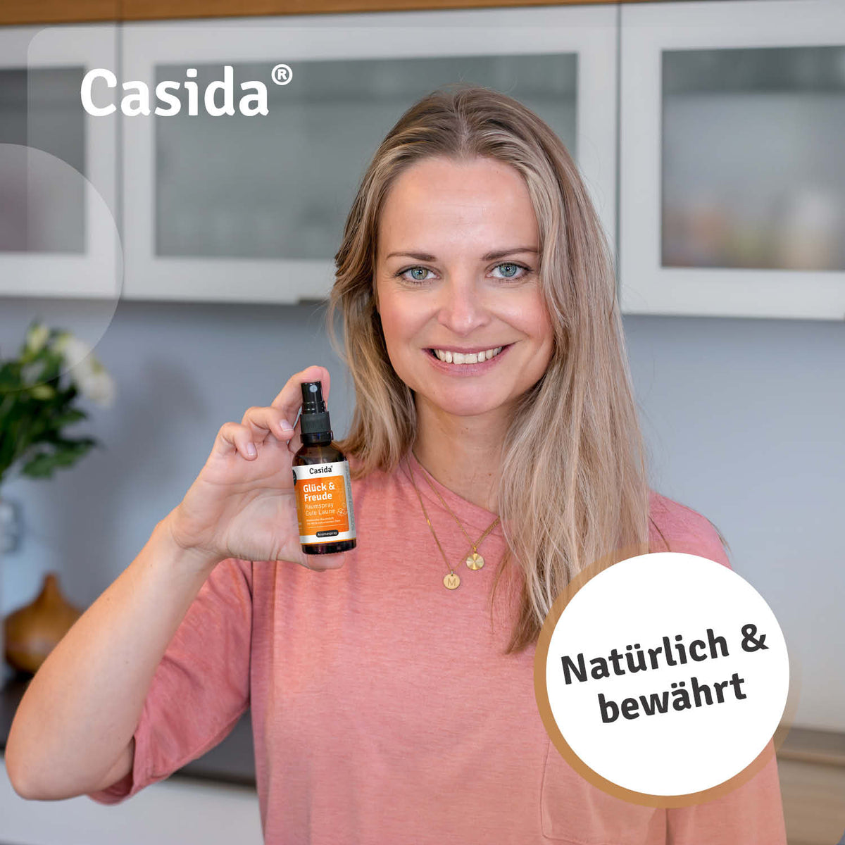 Glück & Freude raumspray held by a woman with long hair, wearing a pink shirt, in a kitchen setting.