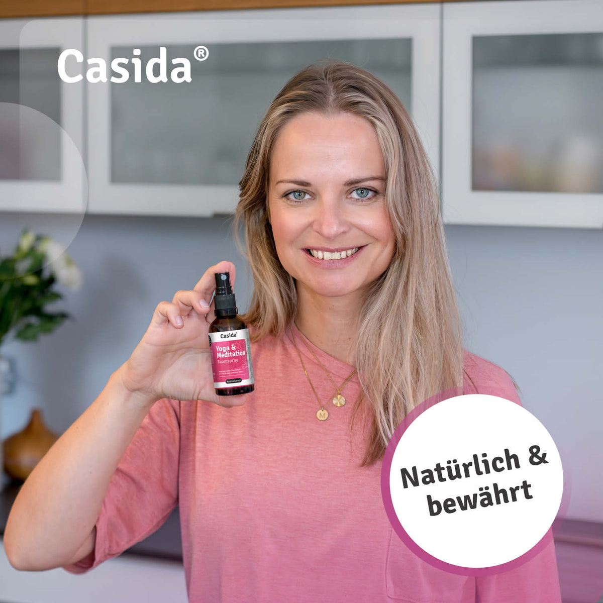Casida yoga and meditation room spray held by a smiling woman in a pink shirt, with a blurred kitchen background.