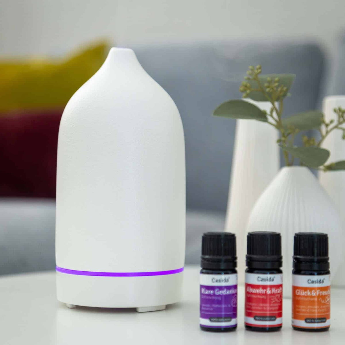 Keramik Aroma Diffuser in white with purple light, accompanied by four essential oil bottles in various colors.