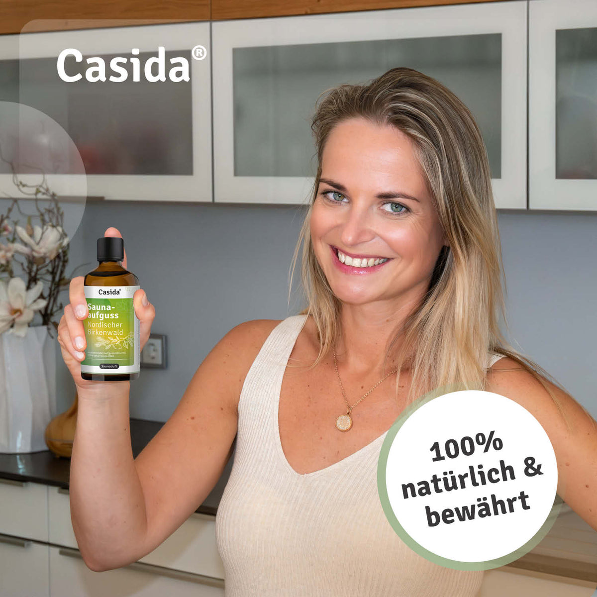Saunaaufguss Nordischer Birkenwald oil in a bottle held by a smiling woman in a modern kitchen setting.